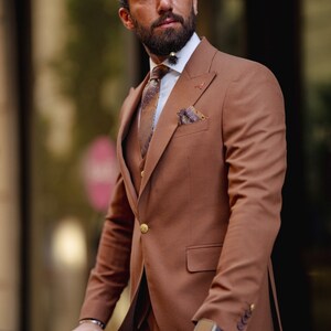 May include: A man wearing a brown suit with a patterned tie and a pocket square. He is standing in front of a window and looking to the side.