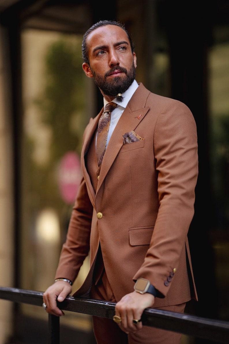 May include: A man wearing a brown suit with a patterned tie and a pocket square. He is standing in front of a window and looking to the side.