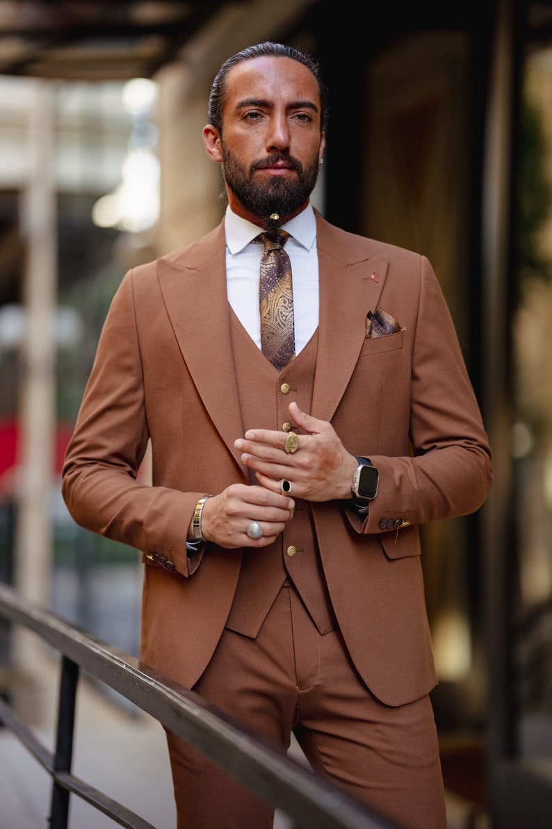 May include: A man wearing a brown three-piece suit with a patterned tie and a pocket square. He is wearing a gold ring on his finger and a watch on his wrist.