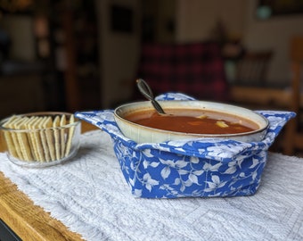 May include: A white bowl with a blue and white floral pattern bowl cozy filled with tomato soup and a spoon. A clear glass bowl filled with crackers sits on a white table cloth.