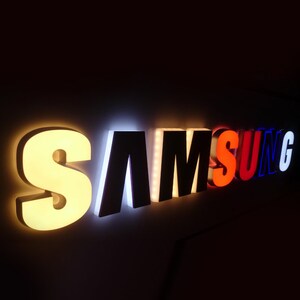 May include: A white, black, red, and blue illuminated sign that reads "SAMSUNG".