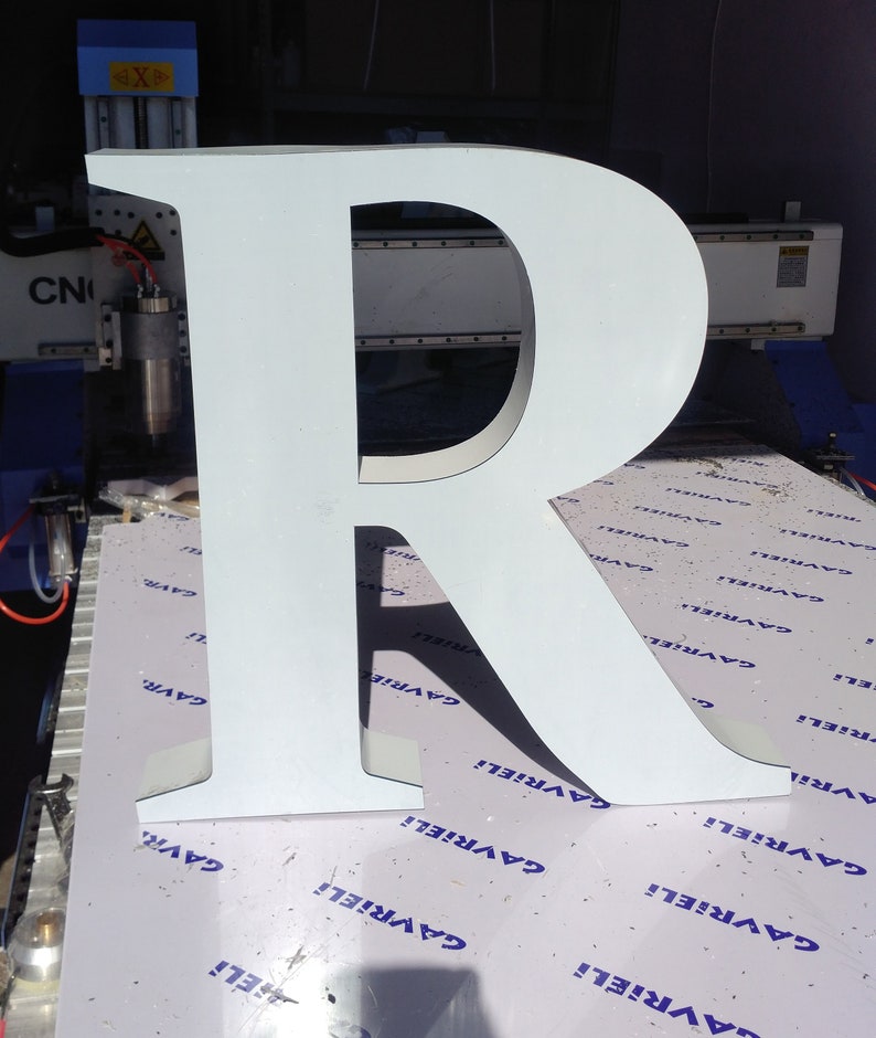 May include: A white, three-dimensional letter "R" being cut out by a CNC machine. The letter is sitting on a white surface with the word "GAVRIELI" printed on it.