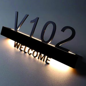 May include: Black and white illuminated house number sign with the number "V102" and the word "WELCOME" in white letters.
