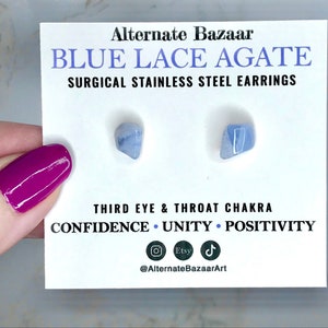 May include: Blue lace agate earrings on a white card with text. The card reads "Blue Lace Agate Surgical Stainless Steel Earrings" and lists "Confidence, Unity, Positivity." The earrings are light blue and the card is held by a hand with magenta nail polish.