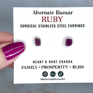 May include: Ruby surgical stainless steel earrings on a white card. The card has the text "Alternate Bazaar" and "RUBY". The earrings are small, rough-cut, and a deep red color. The card also includes the words "HEART & ROOT CHAKRA" and "FAMILY • PROSPERITY • BLISS".