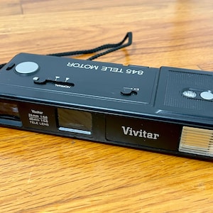 May include: Black Vivitar 845 Tele Motor camera with a lens that reads 24mm f5.6 and 48mm f5.6. The camera has a flash and a black strap. The camera is on a wooden surface.