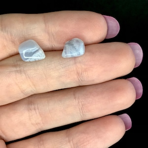 May include: Two small, light blue gemstones with white veining are displayed. The stones are held in a hand against a black background. The gemstones have a smooth, polished appearance.