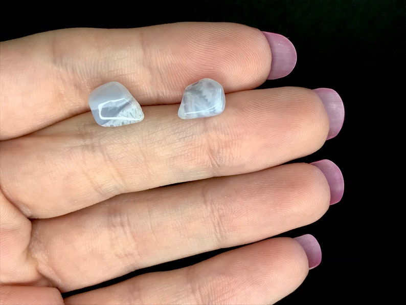 May include: Two small, light blue gemstones with white veining are displayed. The stones are held in a hand against a black background. The gemstones have a smooth, polished appearance.