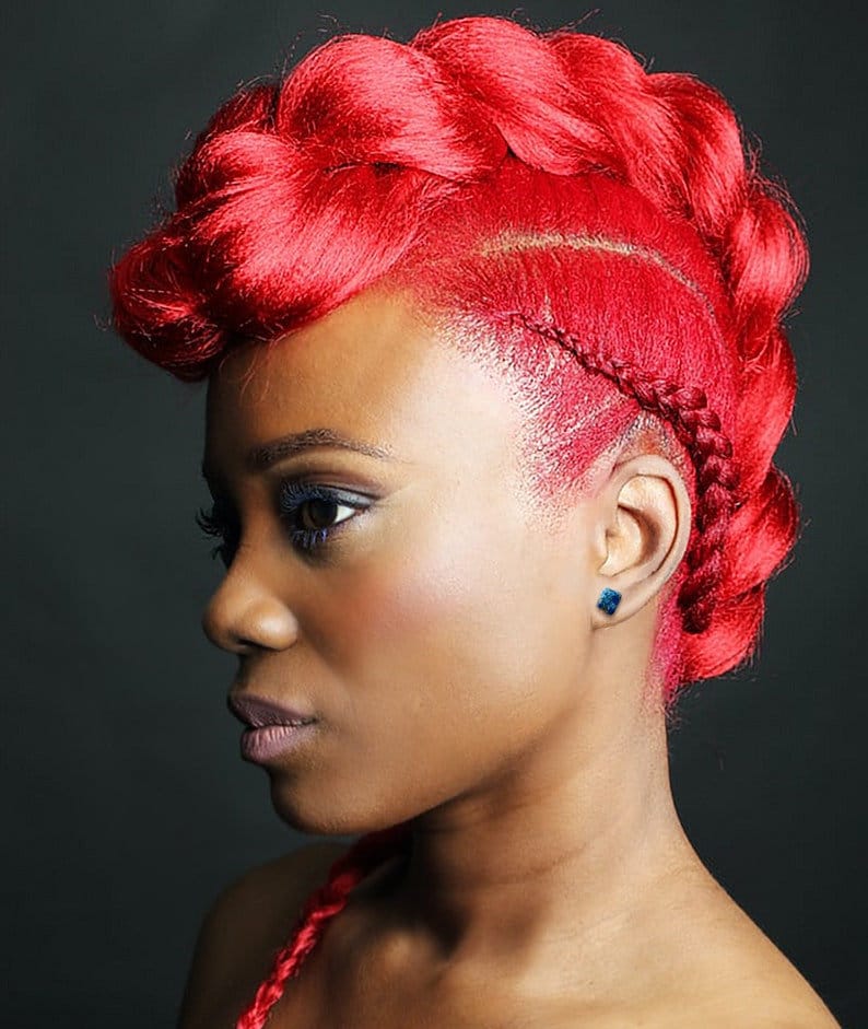 May include: A side profile of a person with vibrant red hair styled in a mohawk-like braid and updo. The hair is a bold, solid red color. The person has dark skin, with blue eyeshadow and a small blue earring.