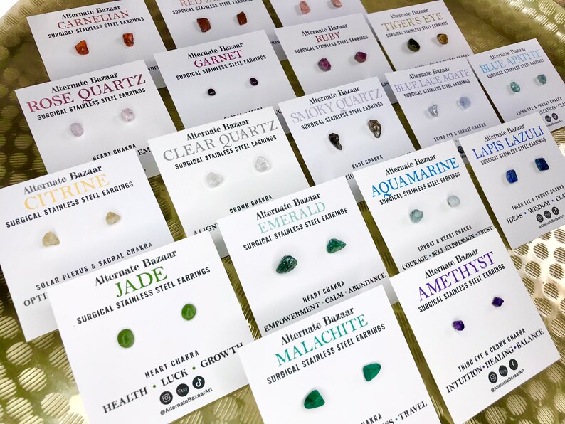 May include: A collection of stud earrings on white cards, each featuring a different gemstone. The cards are labeled with the gemstone name and the phrase "Surgical Stainless Steel Earrings". The gemstones vary in color and shape.