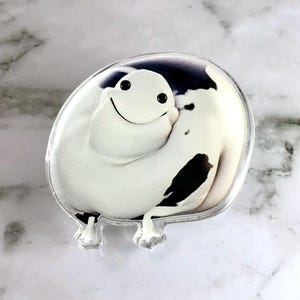 May include: A white and black cartoon character with a big smile, printed on a clear acrylic pin. The character has a round body and a small, black nose.