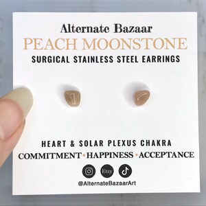 以下が含まれることがあります： 白いカードに、ピーチムーンストーンのスタッドピアスが2つ。カードには「PEACH MOONSTONE SURGICAL STAINLESS STEEL EARRINGS」とチャクラに関するテキストが記載されています。ピアスは小さく、薄茶色で、形が不規則です。