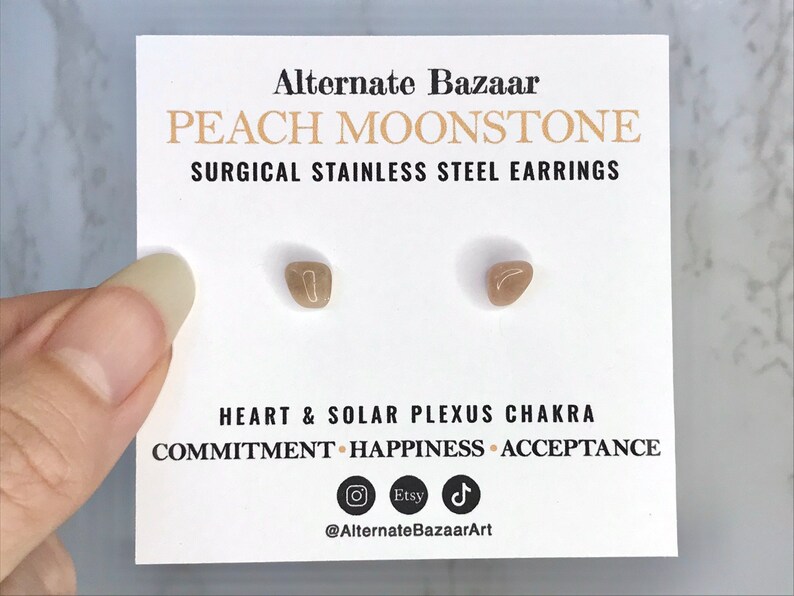 以下が含まれることがあります： 白いカードに、ピーチムーンストーンのスタッドピアスが2つ。カードには「PEACH MOONSTONE SURGICAL STAINLESS STEEL EARRINGS」とチャクラに関するテキストが記載されています。ピアスは小さく、薄茶色で、形が不規則です。