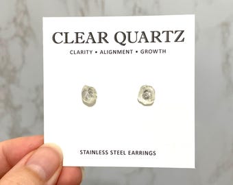 May include: A pair of clear quartz stud earrings mounted on a white display card. The card features the text CLEAR QUARTZ, CLARITY • ALIGNMENT • GROWTH, and STAINLESS STEEL EARRINGS. The earrings are small, irregular, translucent, and colorless.