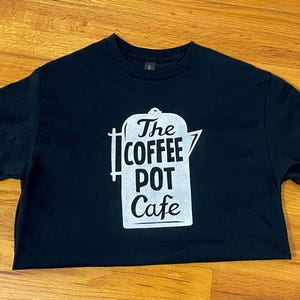 May include: Black t-shirt with a white graphic of a coffee pot and the words "The Coffee Pot Cafe". The shirt is made of a soft material and has short sleeves.