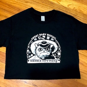 May include: Black t-shirt with a white graphic of a cartoon cat wearing a hat and the text "Sweet Baby Biscuit Marble City Pizza".