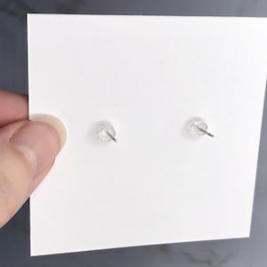 May include: A pair of clear earring backs with metal posts are attached to a white card. The earring backs are made of a soft, clear plastic. The card is held by a hand.