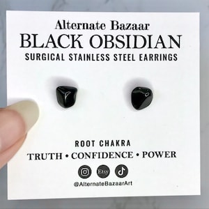 May include: Black obsidian earrings on a white card. The card reads "Alternate Bazaar BLACK OBSIDIAN SURGICAL STAINLESS STEEL EARRINGS." The card also includes the words "ROOT CHAKRA TRUTH CONFIDENCE POWER."