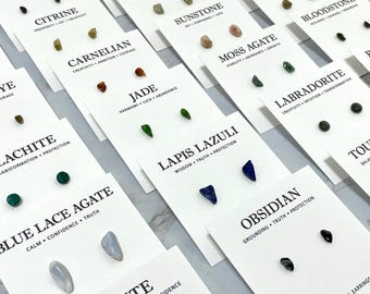 May include: A collection of stud earrings featuring various raw gemstones mounted on white display cards. Each card is labeled with the stone name and associated qualities, including Citrine, Carnelian, Jade, Lapis Lazuli, Obsidian, and Blue Lace Agate.