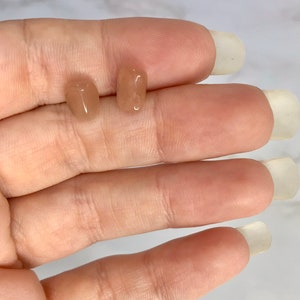 以下が含まれることがあります： 2つの小さく磨かれた、桃色の宝石のイヤリング。1つは楕円形で、もう1つは長方形です。イヤリングは明るい色の表面に表示されています。
