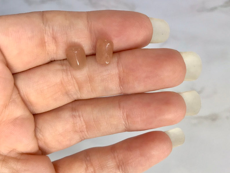 以下が含まれることがあります： 2つの小さく磨かれた、桃色の宝石のイヤリング。1つは楕円形で、もう1つは長方形です。イヤリングは明るい色の表面に表示されています。