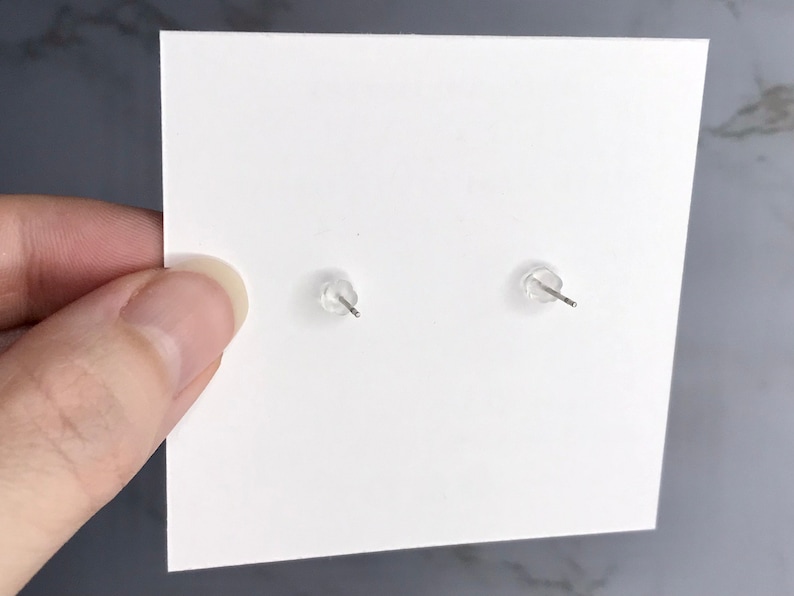 以下が含まれることがあります： 白いカードに、金属製のポストが付いた2つの透明なイヤリングバックが表示されています。 イヤリングバックは透明でゴムのような素材でできています。 カードは手で持たれており、背景はグレーと白です。