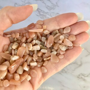 以下が含まれることがあります： 桃色、タン、クリーム色の、磨かれたムーンストーンの宝石の束。石はサイズと形が異なり、滑らかで光沢のある仕上がりです。宝石は自然で素朴な製品です。