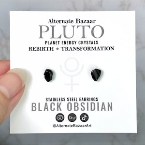May include: A pair of black obsidian stainless steel stud earrings on a white card. The card has the text "Alternate Bazaar PLUTO PLANET ENERGY CRYSTALS REBIRTH TRANSFORMATION BLACK OBSIDIAN". The earrings are small, dark, and irregularly shaped.
