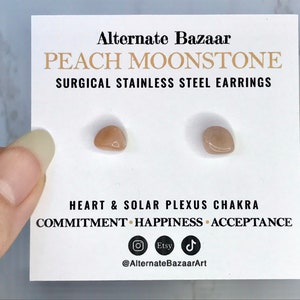以下が含まれることがあります： 白いカードに、ピーチムーンストーンのイヤリングが2つ。カードには「Alternate Bazaar」と「PEACH MOONSTONE SURGICAL STAINLESS STEEL EARRINGS」と書かれています。カードには「HEART & SOLAR PLEXUS CHAKRA COMMITMENT HAPPINESS ACCEPTANCE」も記載されています。