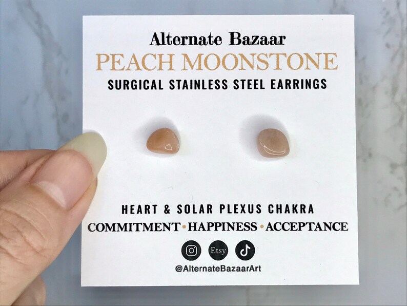 以下が含まれることがあります： 白いカードに、ピーチムーンストーンのイヤリングが2つ。カードには「Alternate Bazaar」と「PEACH MOONSTONE SURGICAL STAINLESS STEEL EARRINGS」と書かれています。カードには「HEART & SOLAR PLEXUS CHAKRA COMMITMENT HAPPINESS ACCEPTANCE」も記載されています。