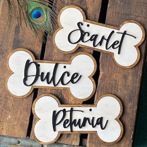 May include: Three wooden dog bone-shaped name tags. Each tag has a light-colored center with a dark wooden border. The names "Scarlet", "Dulce", and "Petunia" are written in a black, cursive font. A peacock feather is in the background.
