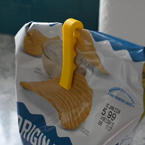 May include: A yellow plastic chip bag clip. The clip is attached to a bag of chips. The bag has a blue and white design with the word "ORIGIN" printed on it.
