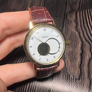 May include: A gold-toned wristwatch with a white face and a brown leather band. The watch face features black numerals, a unique moon phase design, and the word "PAKETA". The band is a dark brown with a crocodile pattern.