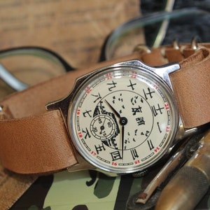 May include: A vintage wristwatch with a light brown leather band. The watch face is cream-colored with black Chinese characters and red numerals. The watch is set against a blurred background with glasses and other items.