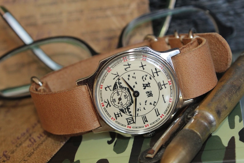 May include: A vintage wristwatch with a light brown leather band. The watch face is cream-colored with black Chinese characters and red numerals. The watch is set against a blurred background with glasses and other items.