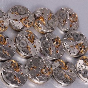 May include: A collection of 12 silver and gold watch movements with intricate gears and details.