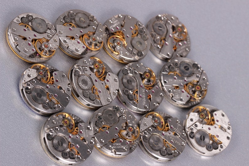 May include: A collection of 12 silver and gold watch movements with intricate gears and details.