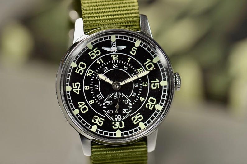 May include: A black and silver wristwatch with a green canvas strap. The watch face has a black background with white markings and a small airplane logo. The watch has a second hand and a 24-hour dial.