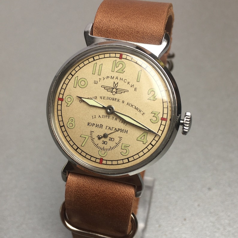 May include: A silver wristwatch with a cream-colored face and black hands. The face has the text "ШТУРМАНСКИЕ" and "ПЕРВЫЙ ЧЕЛОВЕК В КОСМОСЕ" in Cyrillic script. The watch also has the text "12 АПРЕЛЯ 1961" and "ЮРИЙ ГАГАРИН" in Cyrillic script. The watch is on a brown leather strap.
