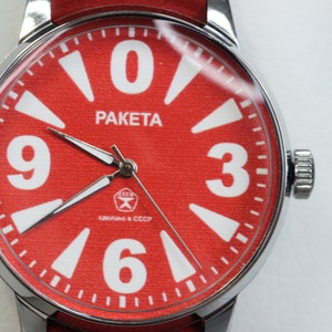 Può includere: Un orologio da polso rosso con numeri e lancette bianchi. Il quadrante dell'orologio dice "PAKEta" e "СССР, сделано в СССР".