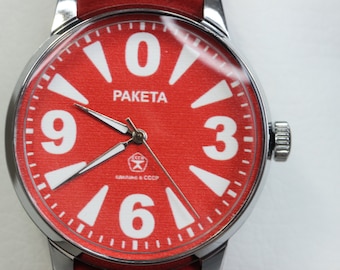 Puede incluir: Un reloj de pulsera rojo con números y manecillas blancas. La esfera del reloj dice "PAKEta" y "СССР, сделано в СССР".