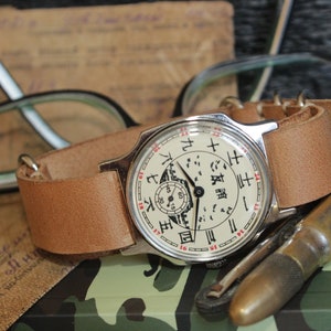 May include: A vintage wristwatch with a light brown leather band. The watch face is off-white with black and red markings, including Chinese characters. The watch is next to eyeglasses, a brass object, and a document.