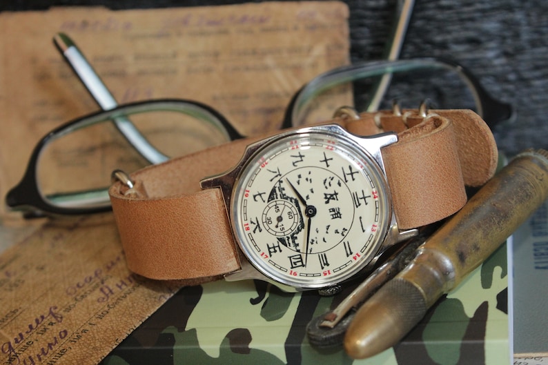 May include: A vintage wristwatch with a light brown leather band. The watch face is off-white with black and red markings, including Chinese characters. The watch is next to eyeglasses, a brass object, and a document.