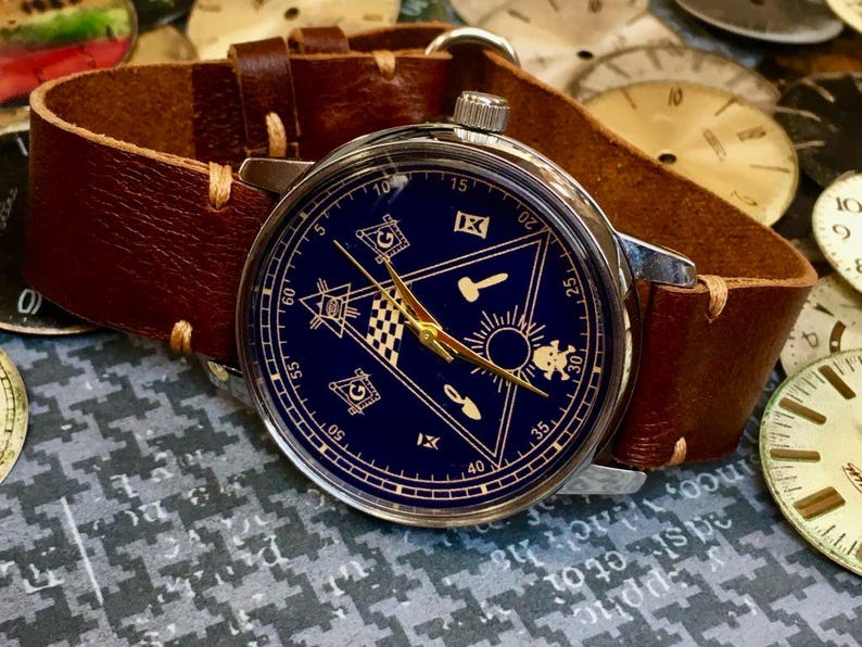 May include: A wristwatch with a dark blue face featuring gold hands and symbols, set in a silver case. The watch has a brown leather band. The watch face displays a triangle with symbols and the numbers 5, 10, 15, 20, 25, 30, 35, 40, 50, and 60.