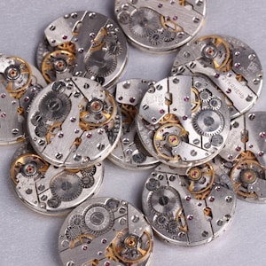 May include: A close-up of a pile of silver watch movements with intricate gears and gold accents. The watch movements are arranged in a random pattern on a silver surface.