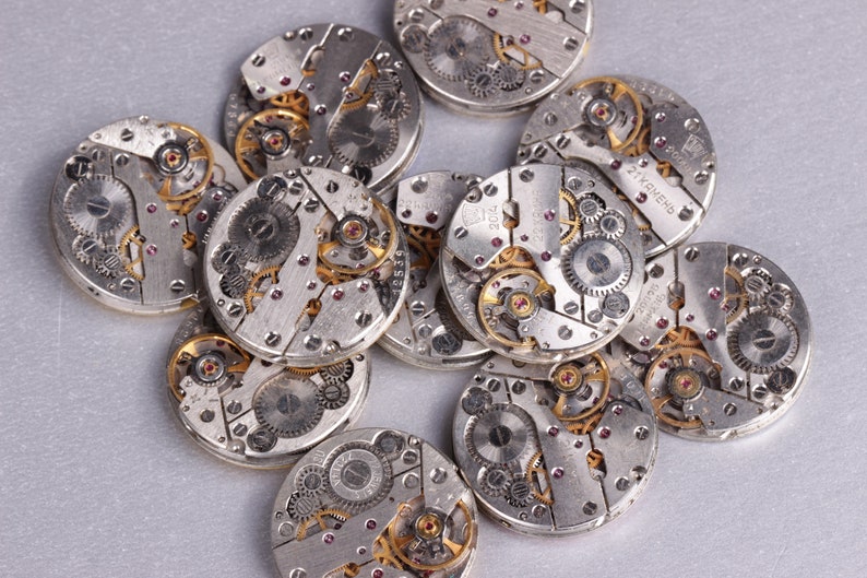May include: A close-up of a pile of silver watch movements with intricate gears and gold accents. The watch movements are arranged in a random pattern on a silver surface.