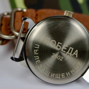 May include: The back of a silver wristwatch with a brown leather band. The watch face has the word "Победа" (Victory) and the number "2602" engraved on it. The text "Пылезащищенные" (Dustproof) is also engraved on the watch face.