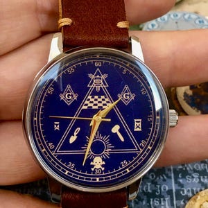 May include: A wristwatch with a dark blue face featuring gold-colored Masonic symbols, including a triangle, square, and compass. The watch has a silver case and a brown leather strap. The watch face also has gold hands and markers.