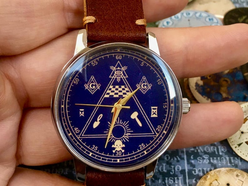May include: A wristwatch with a dark blue face featuring gold-colored Masonic symbols, including a triangle, square, and compass. The watch has a silver case and a brown leather strap. The watch face also has gold hands and markers.