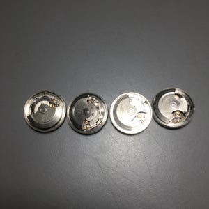 Puede incluir: Se muestran cuatro movimientos de reloj plateados con intrincados mecanismos internos. Las piezas del reloj, de forma circular, revelan engranajes, muelles y otros componentes. La imagen es desde arriba, mostrando la detallada artesan&iacute;a.